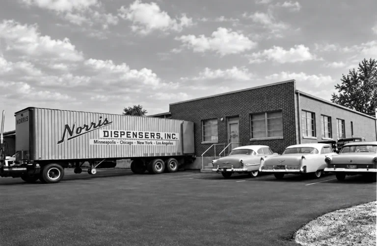 Black-and-white photo of a Norris Dispensers, Inc. trailer and 1950s cars outside a brick building